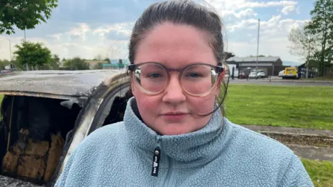 A woman with black, pulled-back hair is standing beside the charred remains of a car. She is wearing black-rimmed glasses and a light blue fleece. She is standing near a large grassy area, with several cars visible in the background.