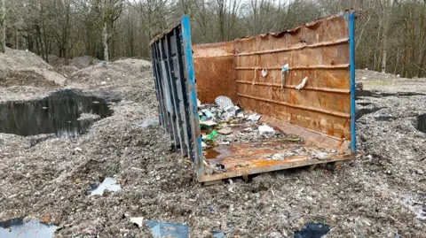 The back of a lorry lies abandoned on piles of waste and water in a woodland clearing