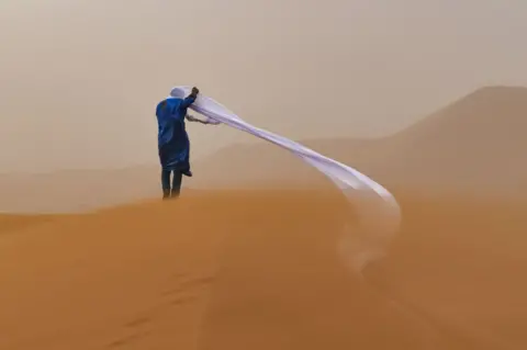 Tom Overall/TNC Photo Contest 2021 A man struggles with wind blowing at cloth around his head whilst standing in the Sahara Desert