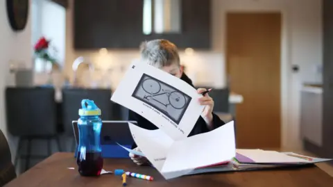 A young boy with short blond hair sitting at a kitchen table on which there is a folder of school work, pens and a drinks bottle. The boy's face is partially obscured as he reads a page of his schoolwork.