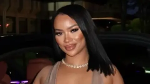 A woman in a light brown dress stands in front of a car, with a shell necklace and straight black hair.