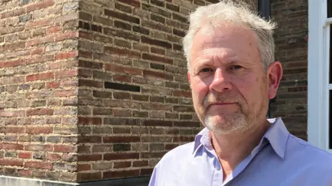 Ed Plowden, who has white hair and a beard, is smiling slightly at the camera. He is wearing an open-necked, light purple shirt. The red brick of City Hall is all that is visible behind him. It is a bright day with the sunlight shining on him and the building.
