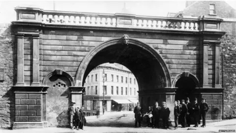 Getty Images A early 20th century black and white photograph of Ferryquay Gate, one of the gates of Derry's walls. An archway can be seen in the old ramparts, through which shops can be seen., A number of men stand at the foot of the gate, as the walls reach up above them