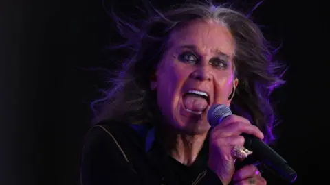 Ozzy Osbourne is holding a microphone with his right hand and has his mouth open. The background is black. 