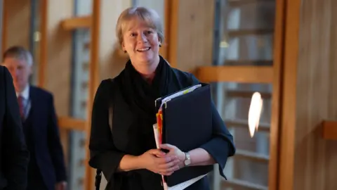 PA Media Shona Robison walking along the corridor at the Scottish Parliament. She has fair hair and is wearing a dark top. She is carrying a black folder, which has sections of paper coming out of the side. She is wearing a light-coloured watch on her left wrist.