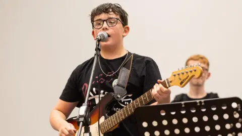 Stepping Forward/Telford & Wrekin Virtual School Matthew, playing a guitar and singing into a microphone, with a person behind him. He is wearing a black T-shirt and has a silver necklace. 