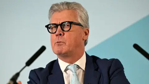 Getty Images Malcolm Offord, who has grey hair and glasses, speaks at a podium. He is wearing a dark suit, white shirt and light blue tie. 