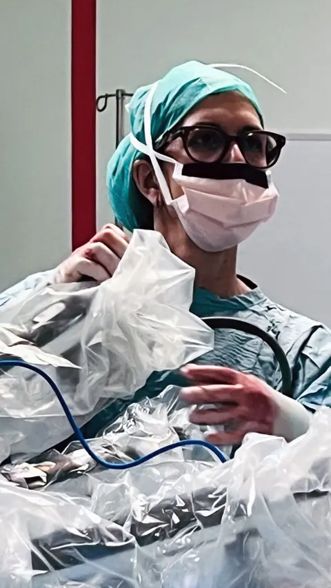 A surgeon in a mask with protective clothing on is holding onto the arms of a surgical robot. She is wearing gloves which are stained red with blood. She has glasses on and is looking away from the robot to the right of the screen.