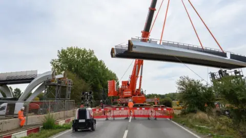 Urban&Civic A large orange crane swinging the flat bridge part of a footbridge into place above the single carriageway A10 near Waterbeach, Cambridgeshire. The road is closed, a worker in an orange hi-vis with a white hat is behind the red barrier. On either side of the road is a verge and hedgerows or trees. On the right is a temporary silver fence and another worker in orange hi-vis with a white hat, sitting on a concrete wall.