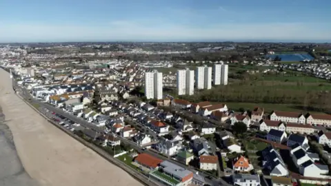 An aerial view of Jersey with the beach in view and the main road followed by rows of houses and four large blocks of flats, blue skies