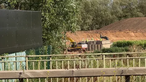 BBC A yellow digger at work in a quarry in the distance. Five people in white hard hats are supervising. There are mounds of soil around the bomb site. Trees are in the background and railings in the foreground