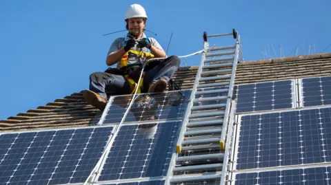 A maintenance engineer wearing a harness and hard hat sits on a roof next to a ladder, servicing solar panels around him on the roof. 