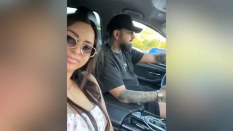 Thames Valley Police A woman and a man in a selfie-style video in the car. The man is driving and the woman is smiling into the camera. They are both wearing sunglasses.