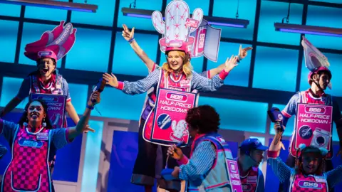 Pamela Raith An image from a musical showing women and men dancing while dressed in bright pink tabards and blue striped shirts. Some have their hands raised in the air while holding pricing guns, while others have pink helmets with cardboard cut-outs attached - one of bananas, one of a cactus, one of a washing machine.  Pink advertising boards on their chests say "half price hoedown" 