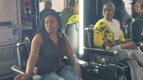 Melanin Moods The two women are sat on barber chairs at the barbers. Chanelle is wearing a black sleeveless top and jeans. Talissa is wearing a yellow cardigan over a white top with jeans. Several men are in the background.