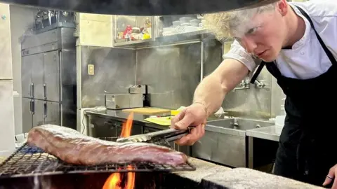 Handout Tommy is cooking a piece of meat over the stove in a kitchen. He is holding a utensil and poking the meat. He has blonde hair and wears a black apron.