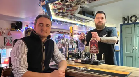 BBC Two men standing either side of a pub bar with beer tapes between them and a green wooden door behind them.