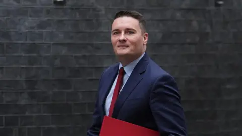 PA Media Wes Streeting, wearing a blue suit and red tie, walks in front of the dark-brick houses of Downing Street while clasping a red file