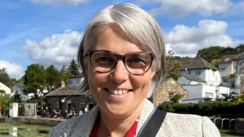 A woman with a grey bob and round glasses, wearing a grey blazer, stands in front of a canal