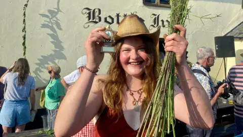 Lucy Dermody, wearing a straw hat, holding a bunch of plucked nettle stems, outside the thatched Bottle Inn pub