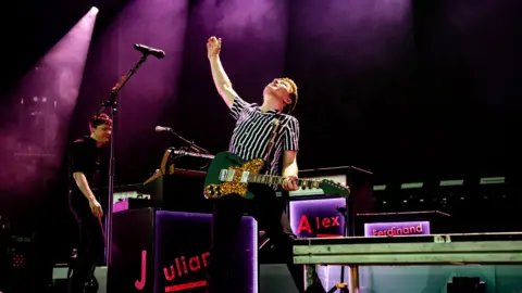 Getty Images Alex Kapranos onstage at a Franz Ferdinand gig,  with one hand wrapped around his guitar and the other raised open-palmed in the air. Bandmate Julian Corrie can be seen playing guitar in the background, and the band's amps are visible too. 