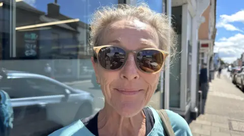 Ben Parker/BBC A woman wearing sunglasses with her hair in a ponytail, she has a blue top on and is looking at the camera. She is stood on a high street.