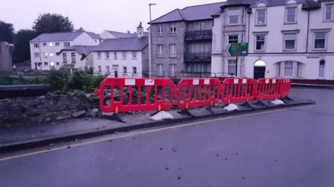 Westmorland and Furness Council A row of plastic fence segments line the path on Nether Bridge. The bridge's parapet wall has collapsed into the river behind it.