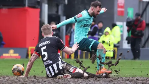Reuters A Grimsby Town player wearing number 5 in black and white stripes slides through thick mud to tackle a Wolves player in a teal kit, with large chunks of mud flying into the air. 