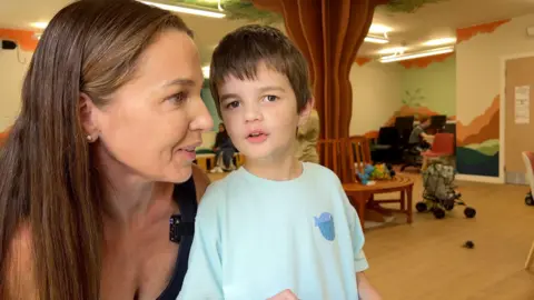Beth is at the side talking to her son Zac. Zac is wearing a blue t-shirt with a whale cartoon on the side of his shirt. 