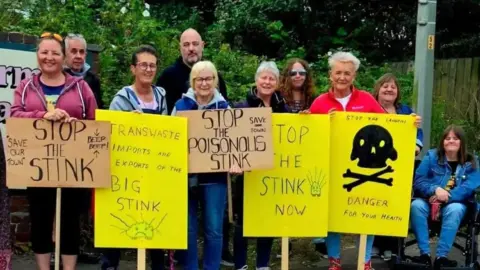 A group of protesters, including MP Lorraine Beavers holding bright yellow placards saying "stop the stink".  There are ten protestors in all, with nine standing behind placards and one lady seated in a wheelchair.