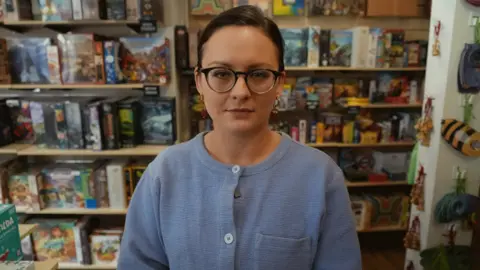 Ms Scaysbrook has dark hair tied up and black rimmed glasses. She wears a light blue buttoned up top. She is inside her shop, with shelves of board games behind her.