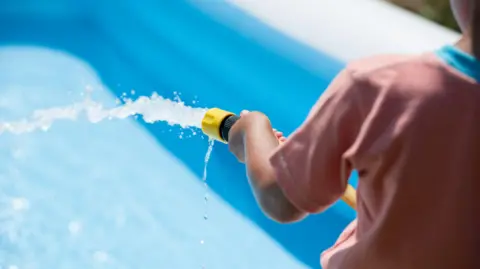 A close-up of a young boy, from behind and wearing a red T-shirt, filling up a blue paddling pool with a hosepipe. 