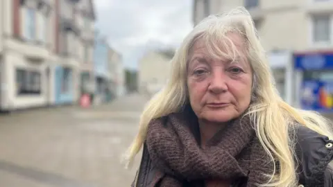 Penny Lloyd wearing a cowel-neck brown sweater and a brown coat and standing in the Triangle in Teignmouth with the street in a blurred background.