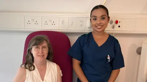 Sarah Ewart is sat in a red chair and stood alongside her is Iru Pyakurel wearing blue scrubs at the Churchill Hospital in Oxford