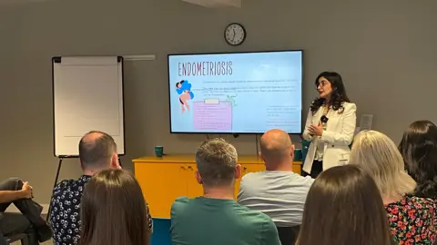 Roya Rasouli, Roya Rasouli is standing in front of a screen which includes information about endometriosis. In front of her are number of people who are sitting down and listing to her talk about the conditon.