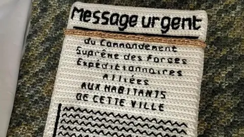 The Longest Yarn A knitted white papered message in French which says 'Message Urgent' and warns residents to leave a town