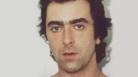 A custody picture of John Cannan. He has very dark wavy hair and dark eyebrows and is looking at the camera with a vacant expression. 