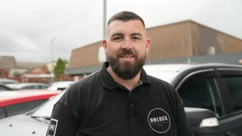 A man with dark hair and a beard is stood in front of a black vehicle. He is wearing a black polo T-shirt and is looking straight into the camera.