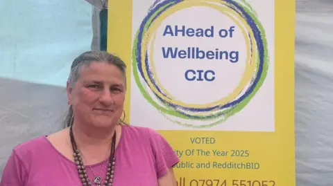 A woman with tied back grey hair wearing a purple t-shirt and brown beaded necklace looks at the camera. She's standing in front of a yellow banner advertising the services and contact number for Ahead of Wellbeing CIC.