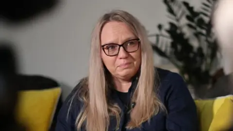 A woman with blonde hair and glasses sits on a settee