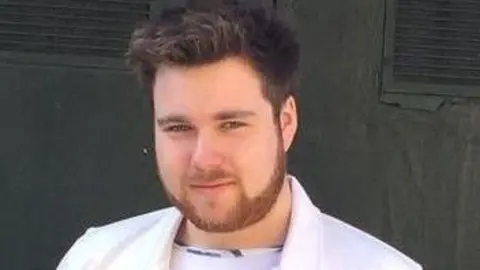South Yorkshire Police Jamie Kelly looks at the camera. He has short, spike brown hair. He is wearing a white jacket and white T-shirt.