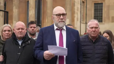 A bald man with glasses, wearing a blue suit, white shirt and purple tie reads a statement outside court flanked by family and friends