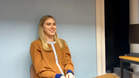 Sadie Reece a young blonde haired girl sat in a chair against a wall smiling.