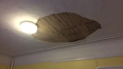 Ison Harrison A hole in a ceiling, showing wooden slates, a light, a white ceiling and yellow walls.