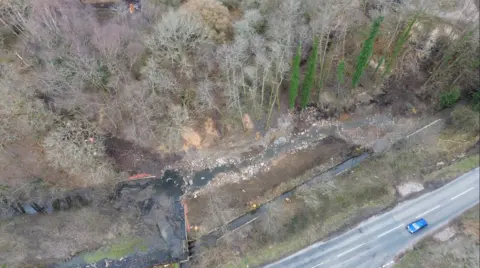 An aerial view of a watercourse by the Forest of Dean, there is a road at the bottom edge and tall green trees by the water bed.