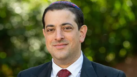 Rabbi Alex Goldberg A man wearing a colourful Jewish skull cap, navy blue blazer and red spotted tie, with a blurred green tree background.