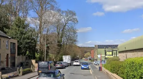 Google A road lined with hedges and houses. In the background is the entrance to a business complex. There are several cars on the road and above is a blue sky, with fluffy clouds.