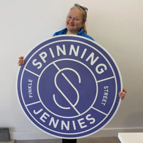 Geri Ward, a blond-haired woman, is standing against a white wall. She has sunglasses on her head and she is smiling. She is holding a circular purple board with the works 'Spinning Jennies Finkle Street' on it.