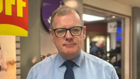 Mr McKillen has short brown hair, black square glasses, a light blue shirt and black tie.