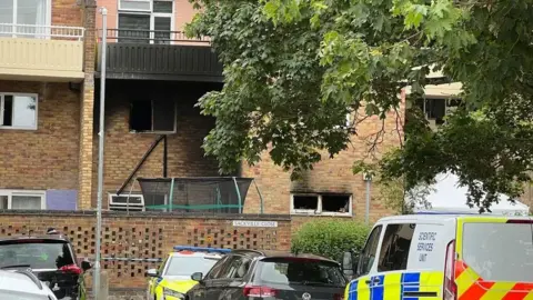 A block of flats is in the background and two of the floors have had a fire which has caused smoke to cling to the bricks. In the foreground there are two police vehicles, and two cars.